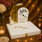 A custom wedding keepsake box set featuring a white arched plaque with gold Arabic calligraphy and names, decorated with white flowers, displayed on top of a white wooden keepsake box with gold lettering. The set is styled on a burlap surface with warm string lights, a brass lantern, and flowers in the background.