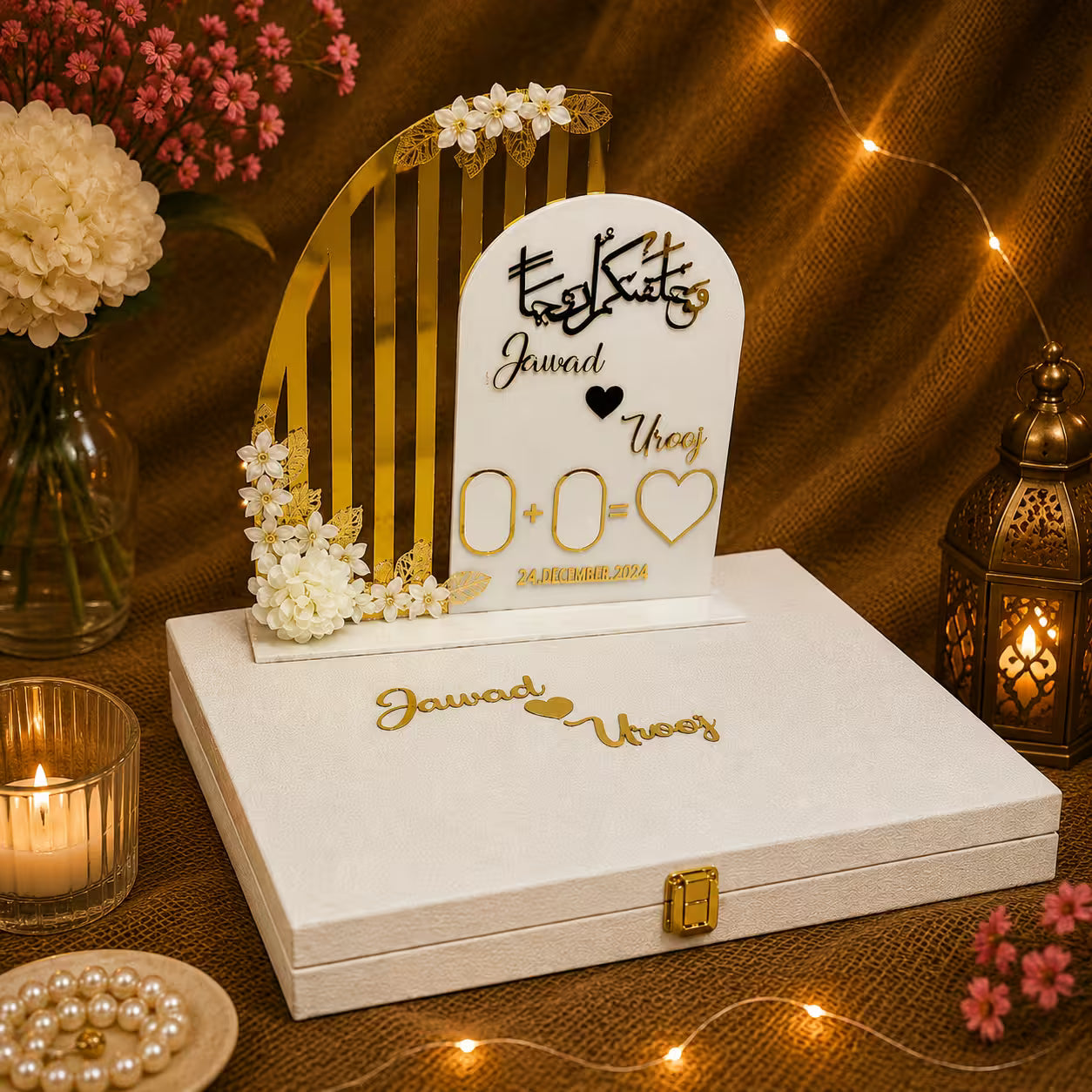 A custom wedding keepsake box set featuring a white arched plaque with gold Arabic calligraphy and names, decorated with white flowers, displayed on top of a white wooden keepsake box with gold lettering. The set is styled on a burlap surface with warm string lights, a brass lantern, and flowers in the background.