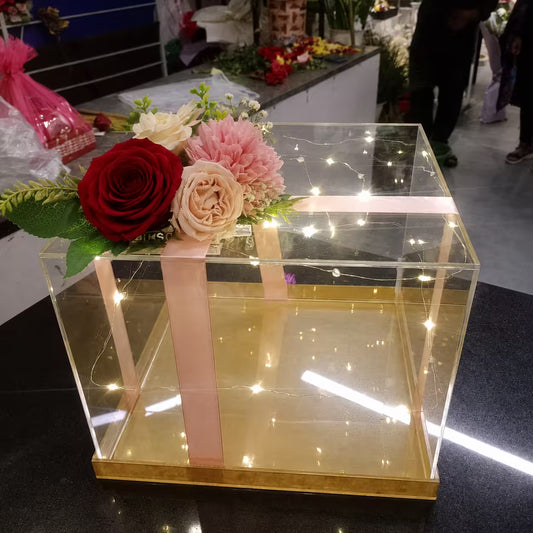 A clear acrylic gift box with a gold base and a pink ribbon, decorated with red and pink artificial flowers on the lid, placed on a dark tabletop in a shop setting.