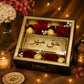 A luxury gift box containing red roses, Ferrero Rocher chocolates, and a gold-toned plaque with Arabic calligraphy reading 'Haq Mehr'. The box is styled on a burlap surface with warm fairy lights, a lit candle, and decorative flowers.