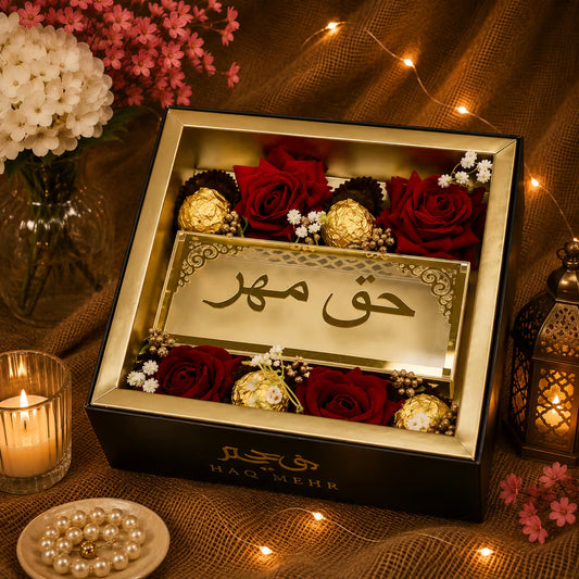 A luxury gift box containing red roses, Ferrero Rocher chocolates, and a gold-toned plaque with Arabic calligraphy reading 'Haq Mehr'. The box is styled on a burlap surface with warm fairy lights, a lit candle, and decorative flowers.