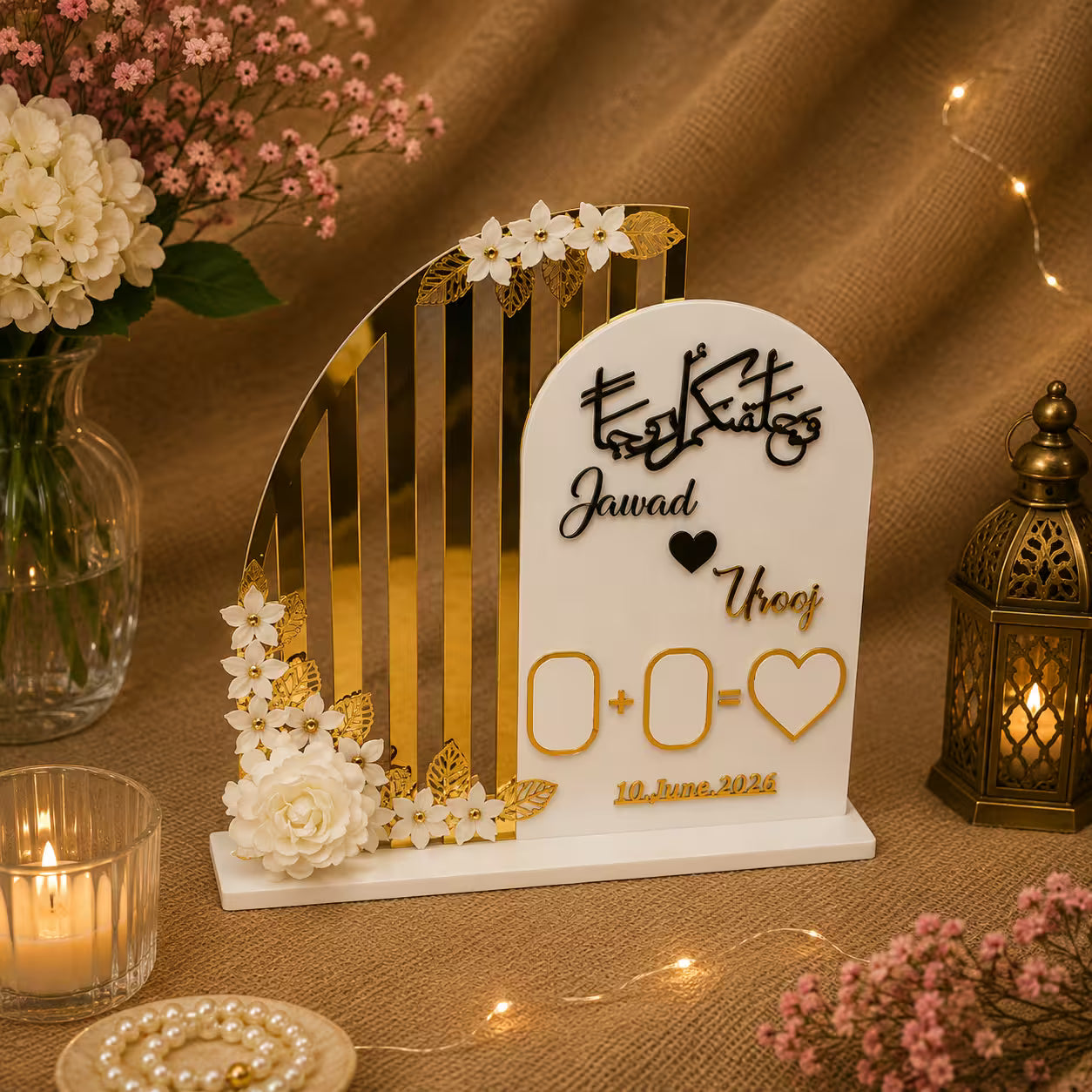 A personalized wedding arch photo board with gold vertical stripes, white floral accents, and custom names 'Jawad' and 'Urooj' on a white arched plaque, styled on a burlap surface with candles, flowers, and a lantern.