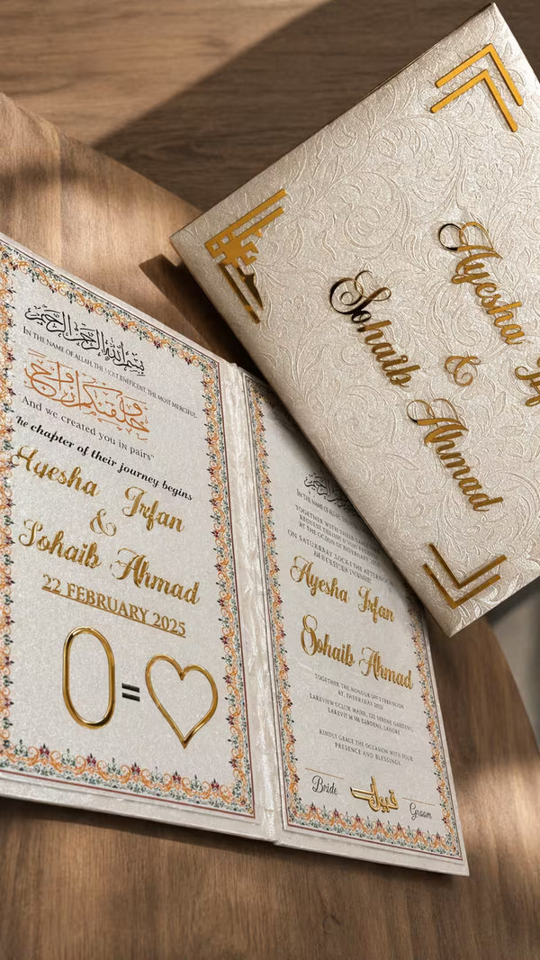 An ornate wedding invitation suite displayed on a wooden surface, featuring a textured cream envelope with gold foil calligraphy and a matching open invitation card with gold decorative borders and text.
