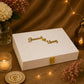 A white textured fabric keepsake box with a gold clasp and personalized gold script reading 'Jawad loves Uooy' on top, styled on a burlap surface with warm string lights, flowers, and candles.