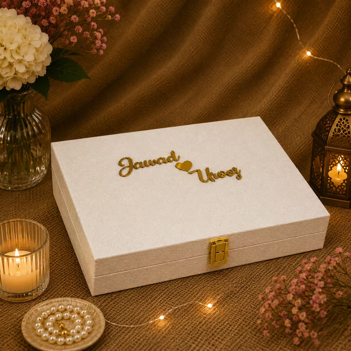 A white textured fabric keepsake box with a gold clasp and personalized gold script reading 'Jawad loves Uooy' on top, styled on a burlap surface with warm string lights, flowers, and candles.