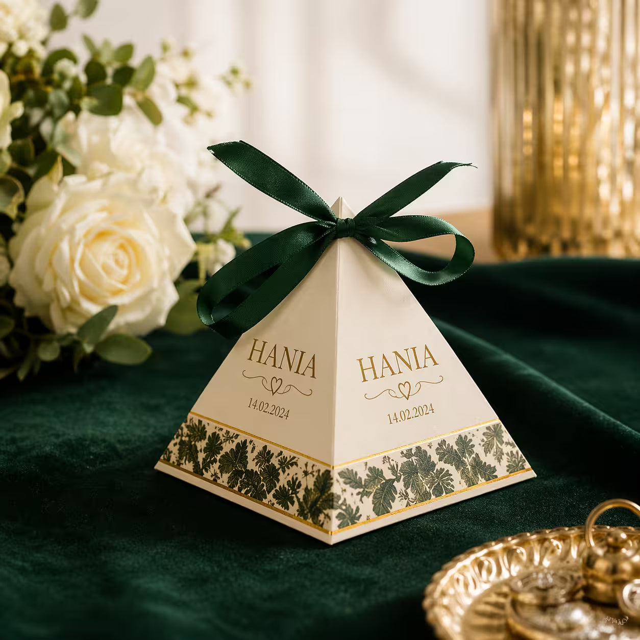 A personalized pyramid-shaped favor box with a dark green ribbon bow, placed on a dark green velvet surface with white roses and gold decor in the background.