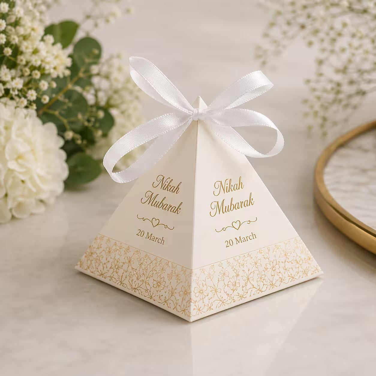 A square pyramid wedding favor box with a white satin ribbon bow, personalized with gold foil text reading 'Nikah Mubarak' and the date '20 March', set on a light marble surface with white flowers in the background.